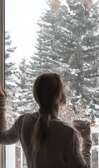 Donna osserva la neve da una finestra in alluminio con isolamento termico – comfort e protezione con Aluminia